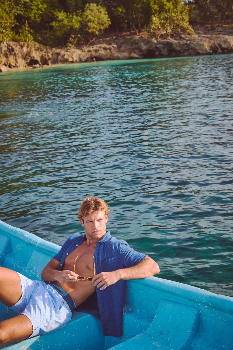 A blond adult male in shorts and an unbuttoned shirt reclines in a blue boat on blue water with a forested shoreline in the background.