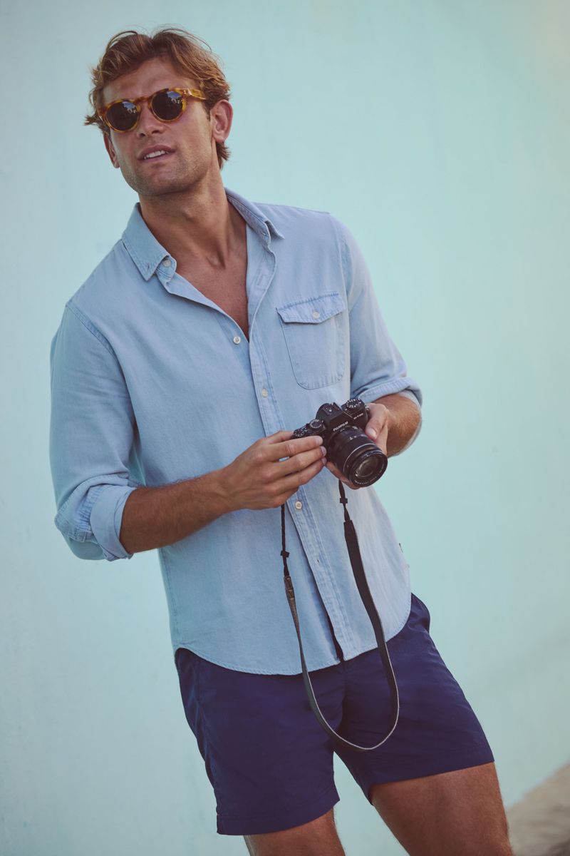 Man in light blue shirt and navy shorts holding black camera with strap in front of light blue wall. Sunglasses, looking left.