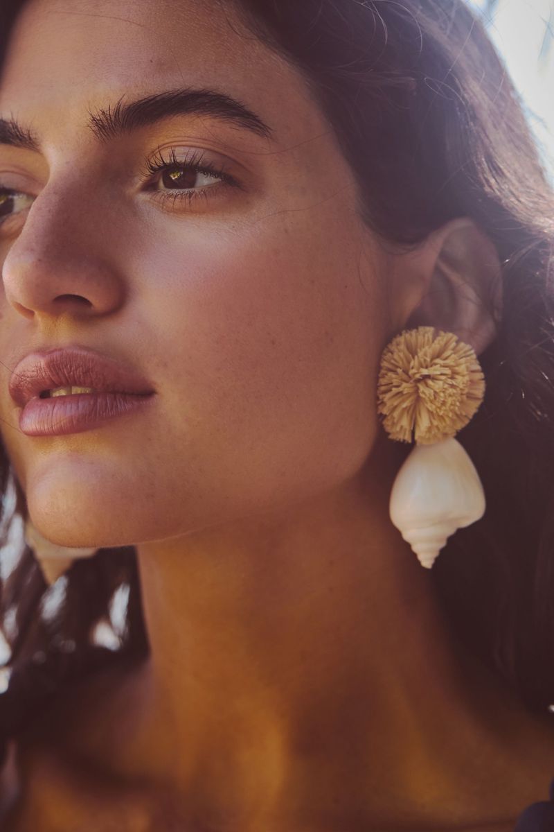Close-up of a woman with dark wavy hair wearing statement earrings, featuring a straw pom-pom and a large shell.
