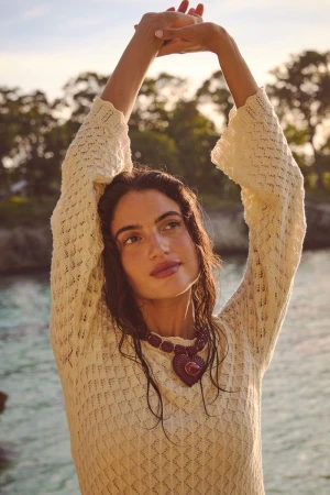 Model Rafaella Consentino with arms raised overhead wearing a cream open-knit sweater and heart pendant necklace outdoors.