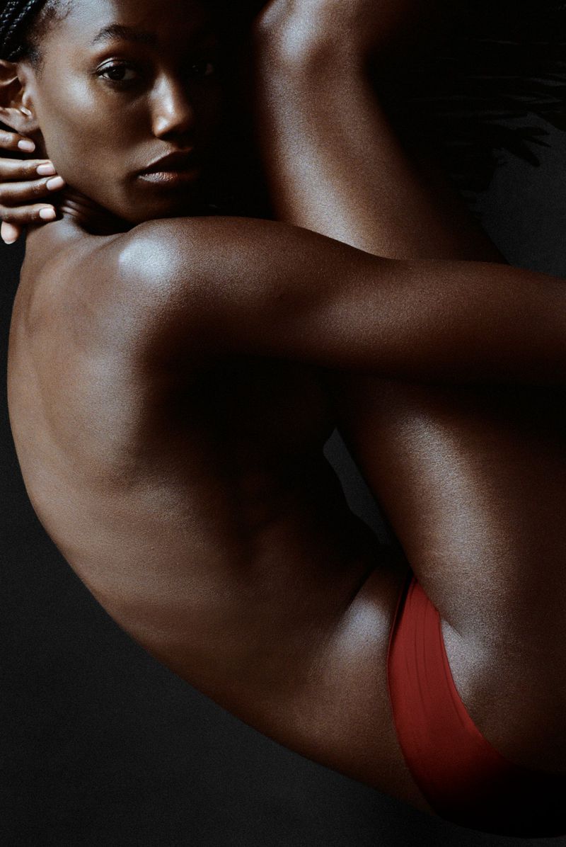 Striking studio shot of a beautiful adult Black woman embracing her legs, highlighting her skin tone and minimalist red undergarment.