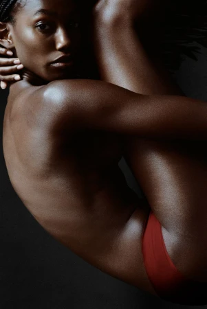 Striking studio shot of a beautiful adult Black woman embracing her legs, highlighting her skin tone and minimalist red undergarment.