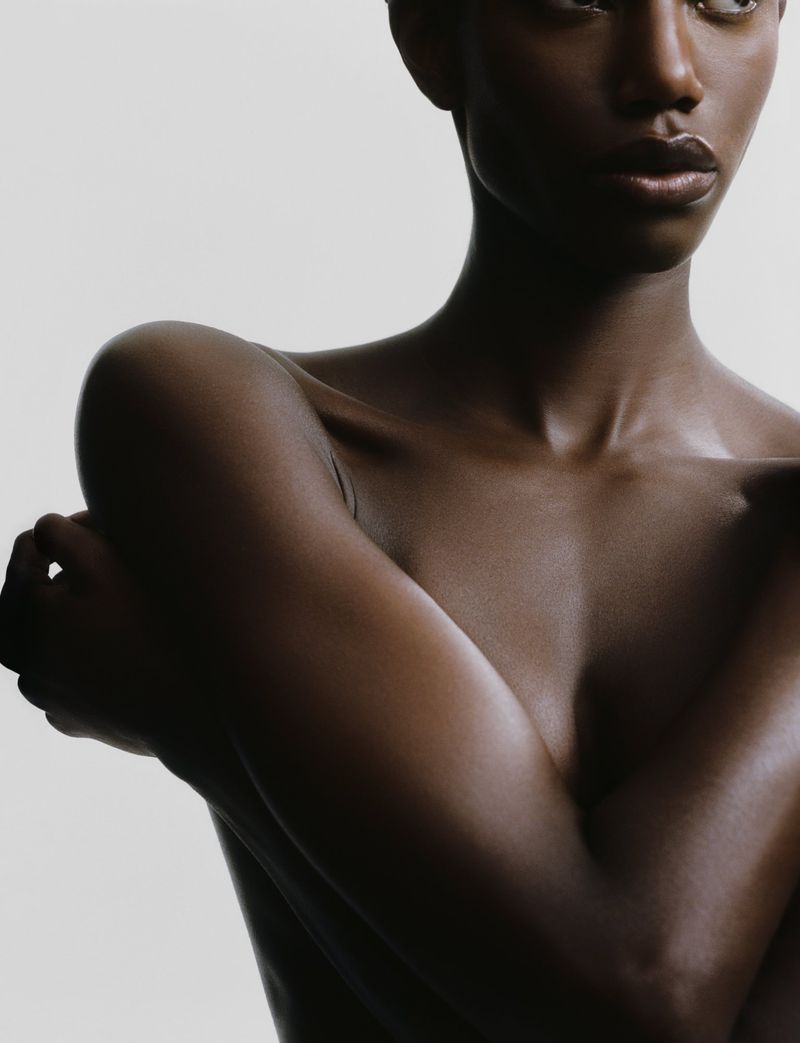 Close-up studio shot of a dark-skinned woman with arms crossed, showcasing the shoulders and upper chest against a plain background.