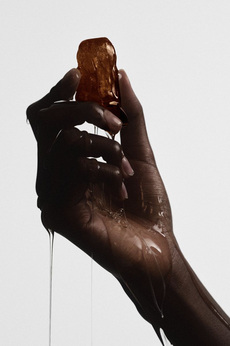 A dark-skinned hand holds an amber cube from which liquid drips, against a light background, creating a visually arresting beauty shot.