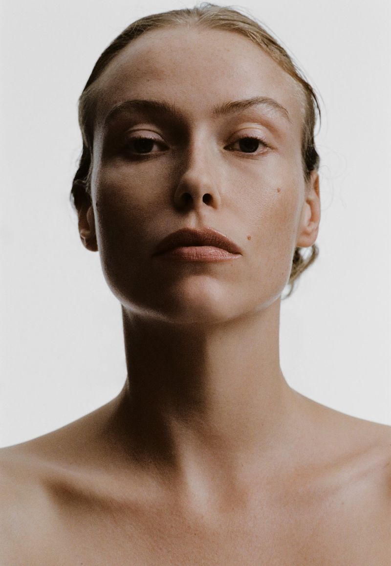 Close-up portrait of a fair-skinned woman with slicked-back blonde hair against a light background, showcasing natural beauty and neutral tones.
