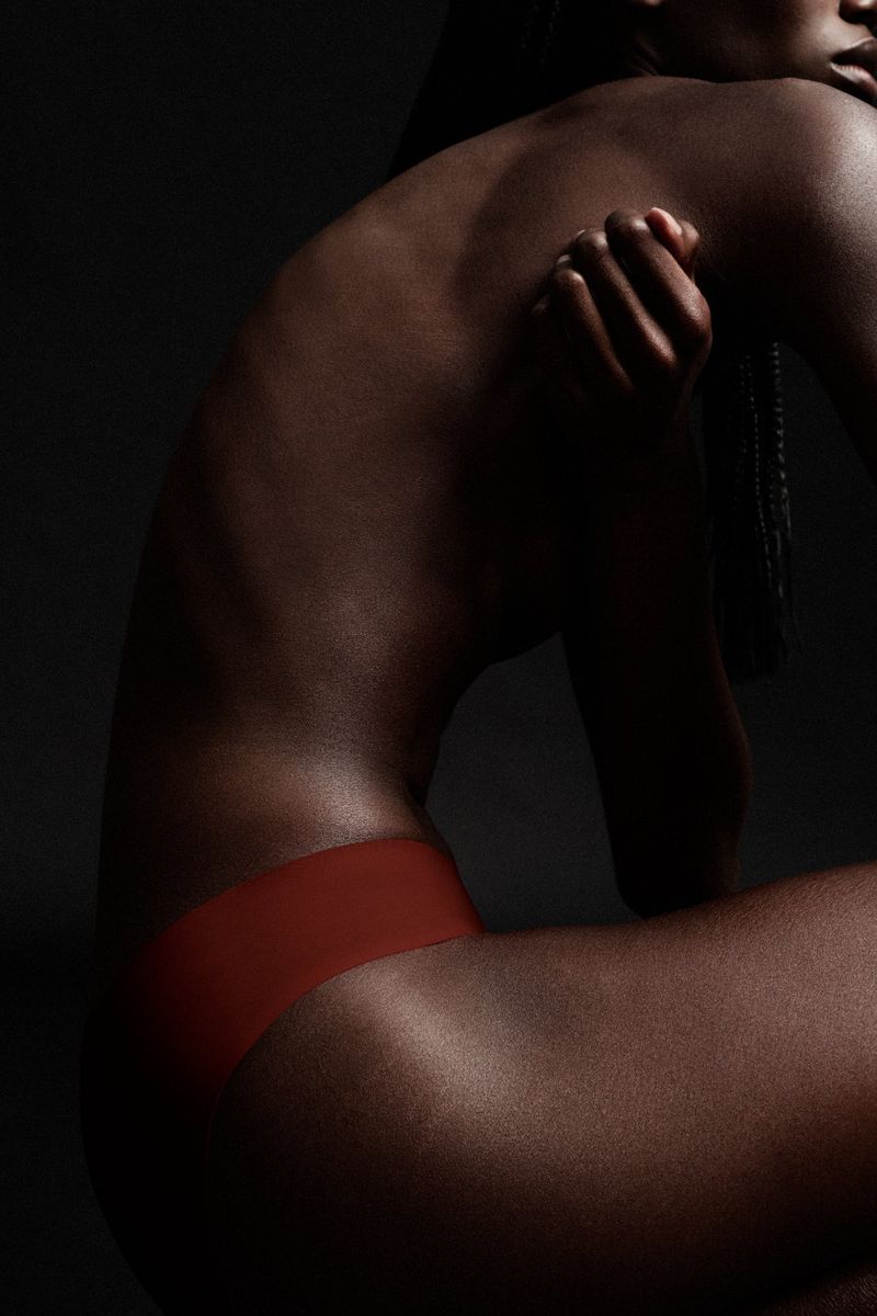 Striking studio shot of a Black female model with bare back and minimalist bottoms against a dark background, showcasing natural beauty and form.