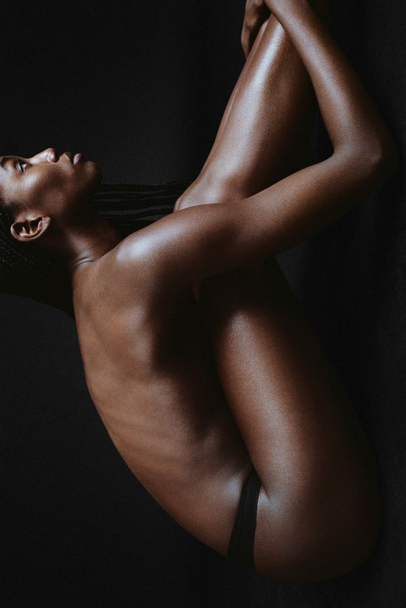 A dramatic close-up studio shot of a Black adult female model in minimal attire, emphasizing her pose against a stark black background.
