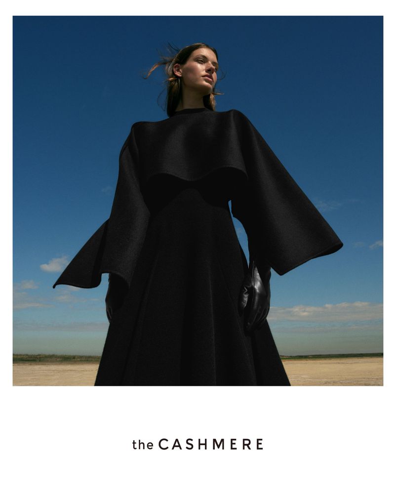 Woman in flowing black dress stands against a blue sky and neutral earth tones in a fashion editorial shot with natural lighting.