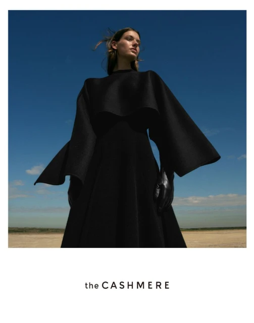 Woman in flowing black dress stands against a blue sky and neutral earth tones in a fashion editorial shot with natural lighting.