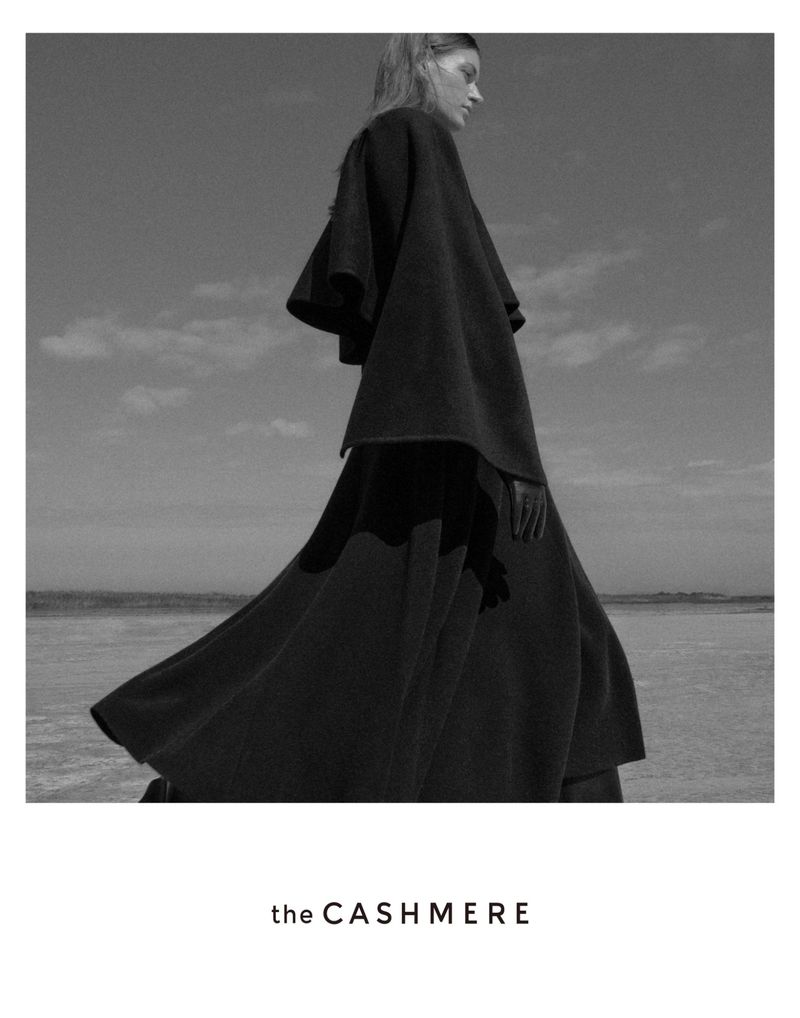 Black and white fashion shot of a woman in a cape-like garment, on a cloudy day by the ocean, styled for a high-fashion editorial.