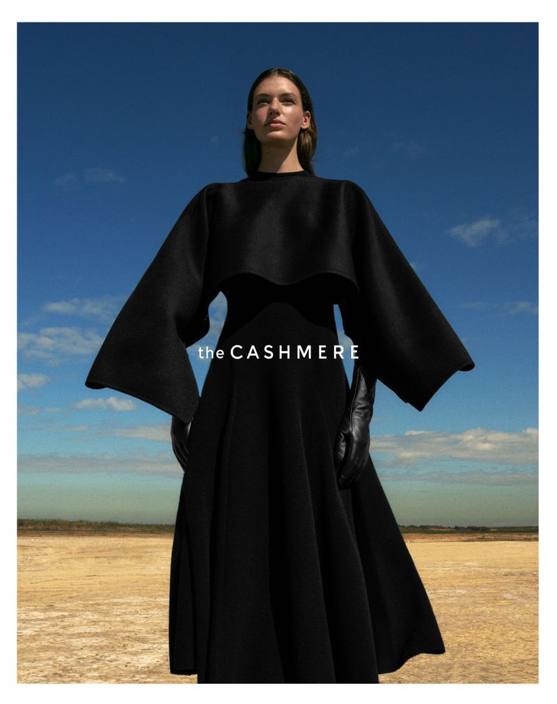 Elegant woman in black dress with cape posing outdoors under blue sky and clouds. Fashion editorial shot with dramatic lighting.