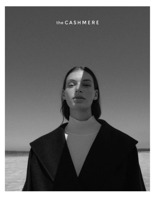 Black and white portrait of a woman on the beach wearing a high-necked white shirt and a dark coat against a clear sky.