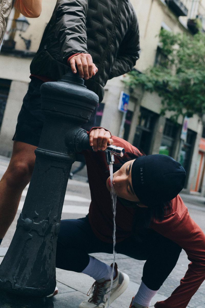 Woman drinks from outdoor public fountain in urban setting, wearing athletic wear, with another person visible in background.