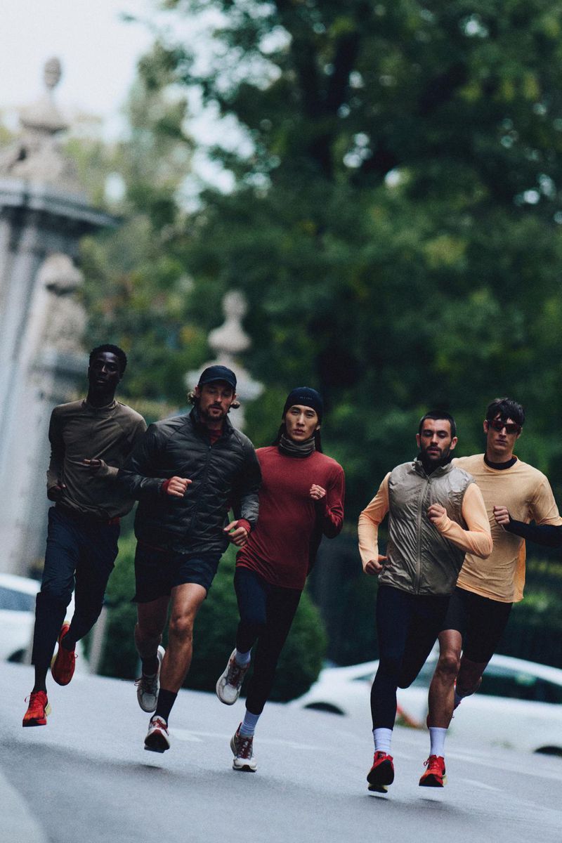 Five diverse male athletes run outdoors on a city street wearing athletic wear, showcasing speed and fitness against a blurred cityscape backdrop.