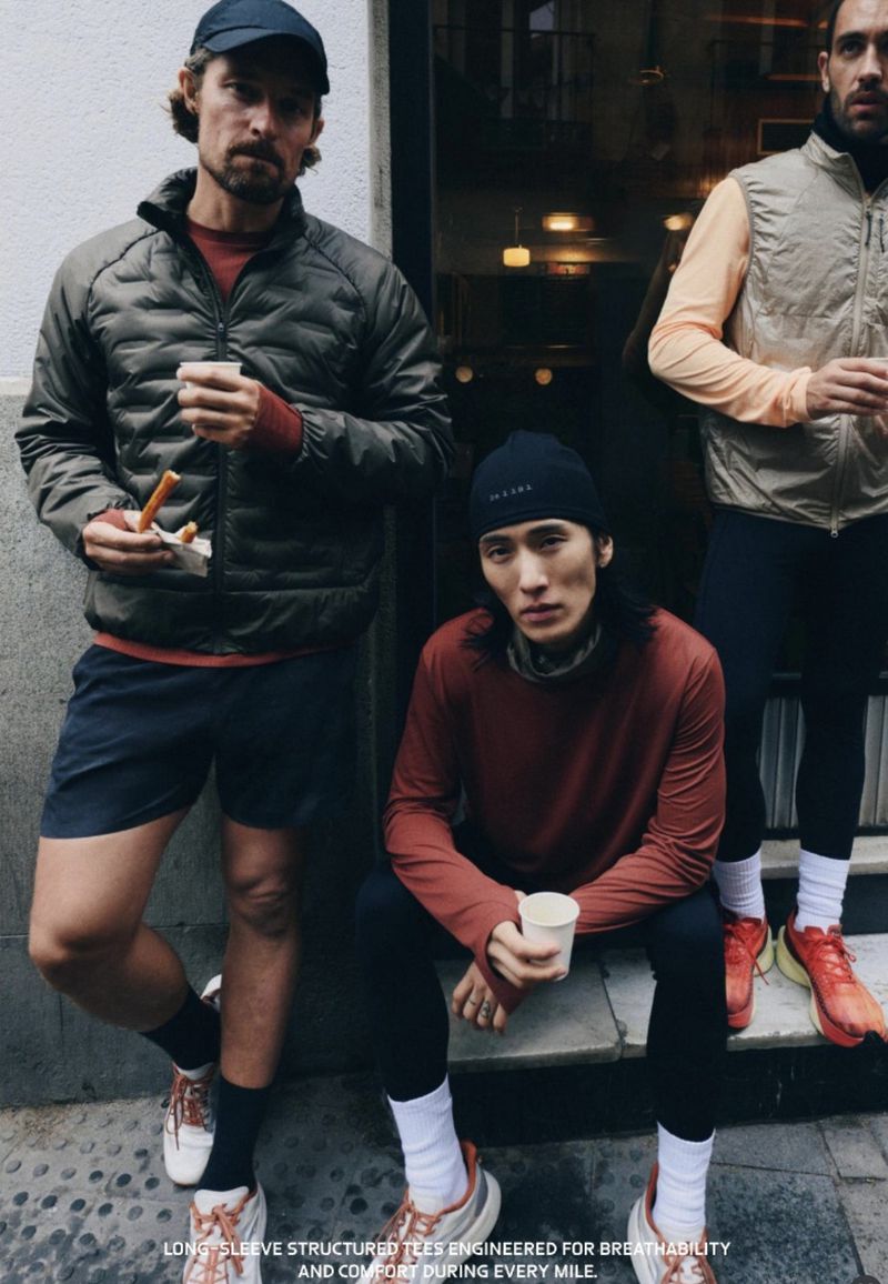 Three people in athletic wear stand and sit outdoors in an urban setting, holding cups and snacks, in a medium shot with natural light.