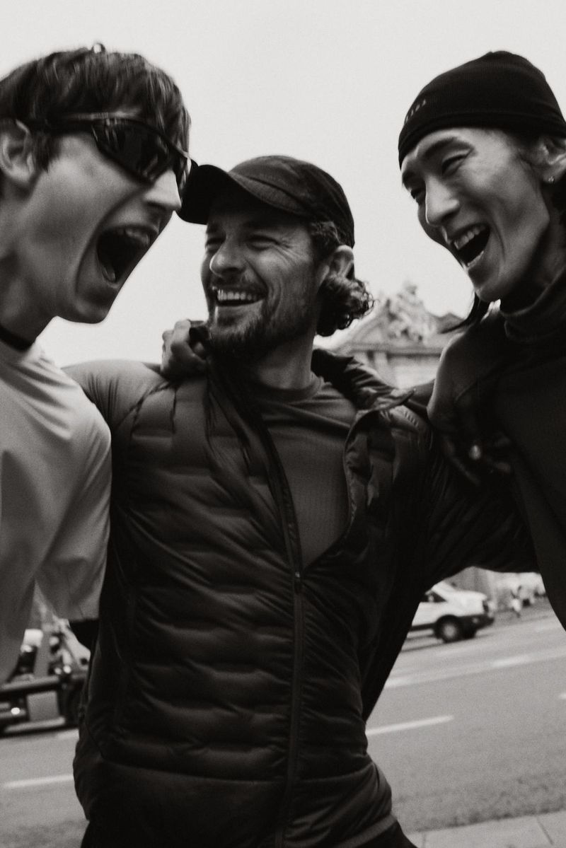 Three smiling adults embrace outdoors on a street, wearing a hat, beanie, and sunglasses, showcasing joy and camaraderie in monochrome.