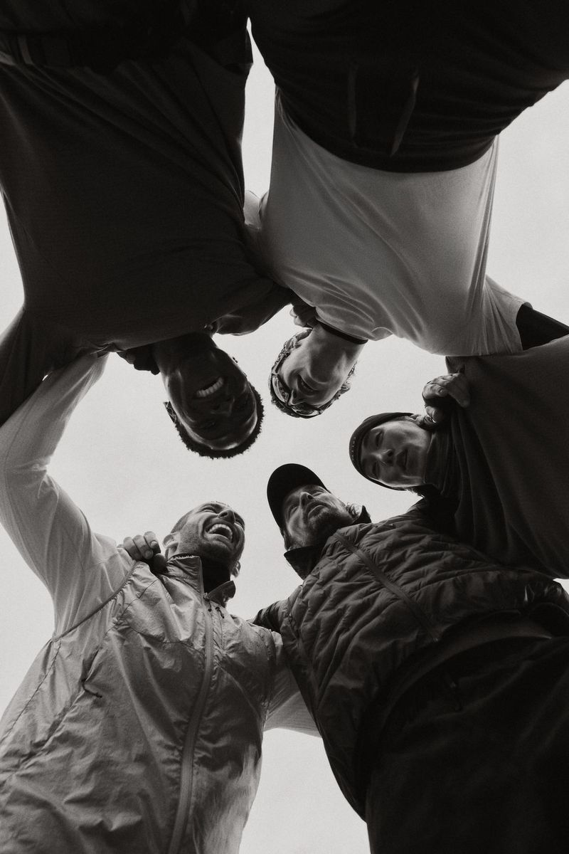 Group of five people huddle together smiling, captured from a low angle looking up, in black and white tones, creating a sense of unity.