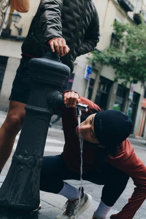 Model wearing a black beanie and red jacket crouching to drink water from a street fountain in the city