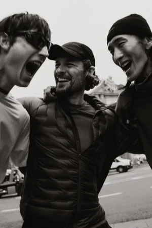 Three men laughing and shouting together outdoors on a city street wearing jackets, beanies, and caps in a black and white shot.