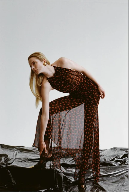 A woman poses in a long, sheer brown and red polka dot dress, leaning forward to adjust her black high heels against a white backdrop.
