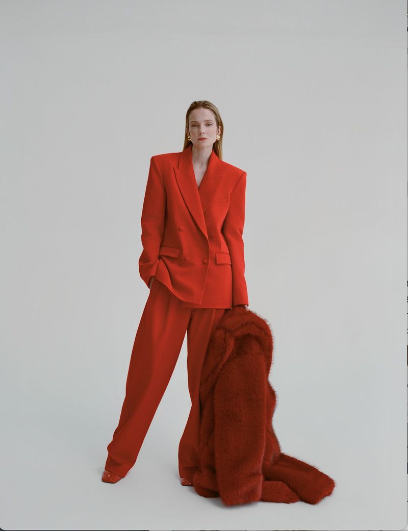 A stylish woman in a red suit poses in a studio, holding a red faux fur coat. The image features clean lines and a minimalist background.