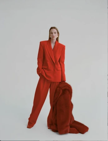A stylish woman in a red suit poses in a studio, holding a red faux fur coat. The image features clean lines and a minimalist background.