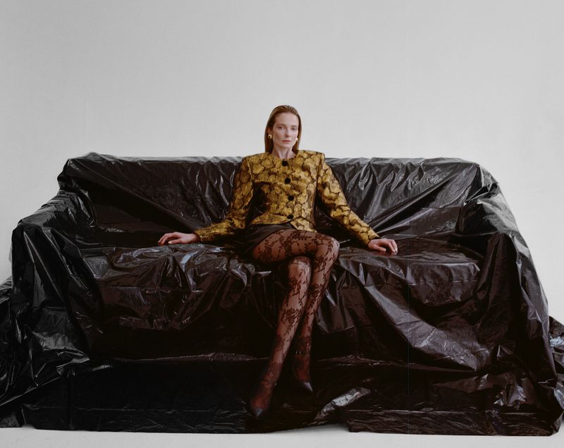 Woman in a floral jacket and black lace tights poses on a couch covered in black plastic sheeting in a brightly lit studio.
