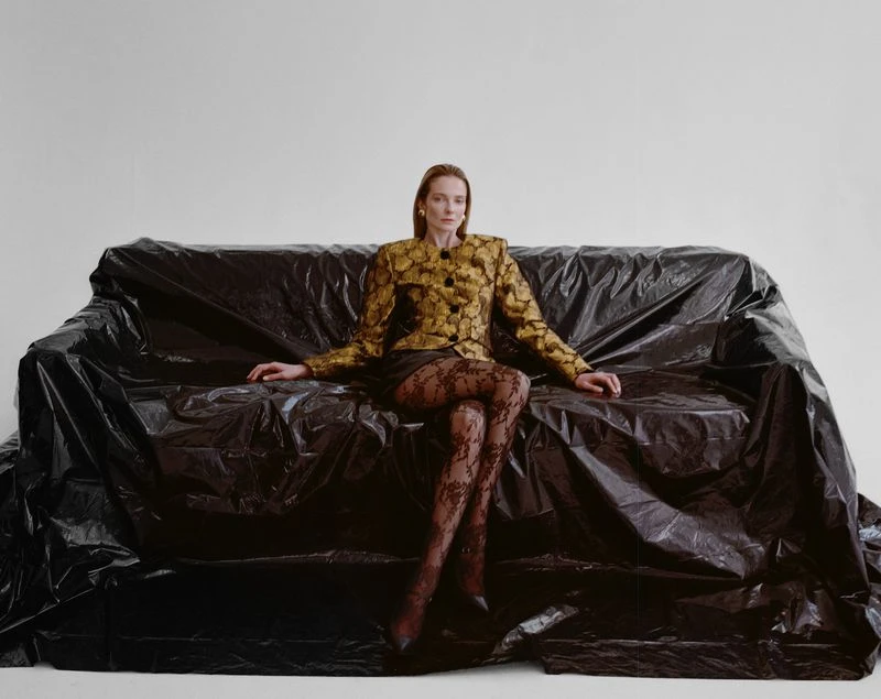 Woman in a floral jacket and black lace tights poses on a couch covered in black plastic sheeting in a brightly lit studio.