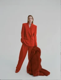 Eniko Mihalik wearing a vibrant red suit holding a matching furry coat against a plain white studio backdrop.