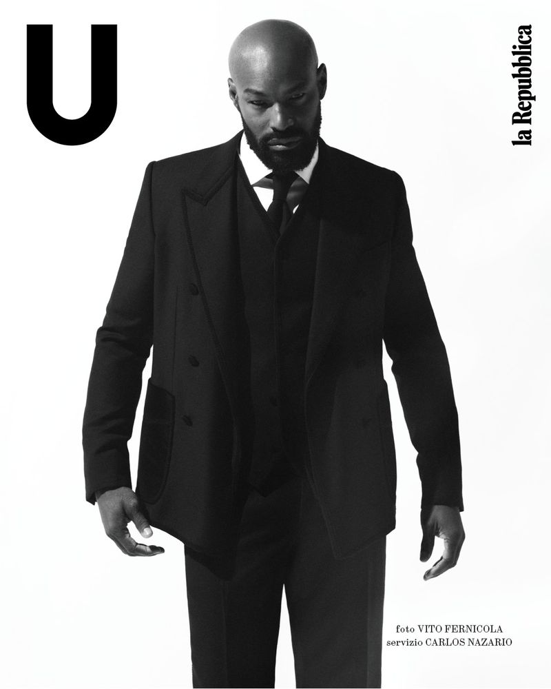 Black and white fashion portrait of a bald African American man in a dark suit, standing against a bright background, looking down.
