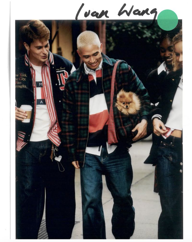 Three people in 90s fashion including plaid jacket, jeans, varsity jacket, and a dog in a shoulder bag on an outdoor walkway.