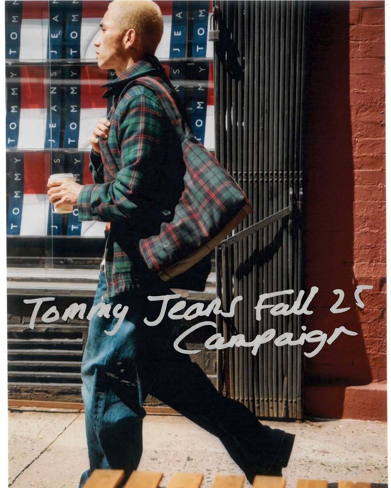 Adult male in outdoor fashion shot. Man walks past the Tommy Jeans store display, wearing a flannel shirt and jeans, with a messenger bag.