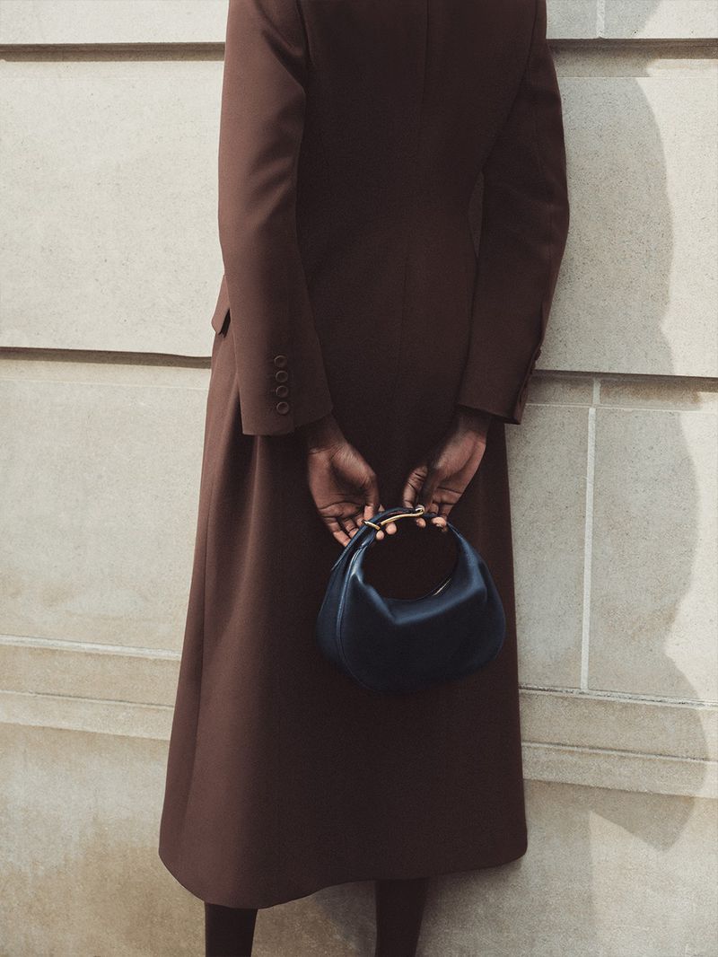 Woman in brown coat holds dark handbag with gold handle against light wall in fashion photography. Minimalist style, neutral palette.