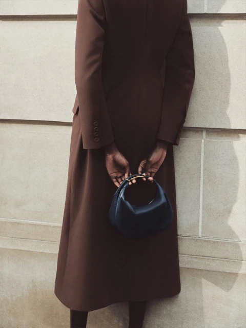 Woman in brown coat holds dark handbag with gold handle against light wall in fashion photography. Minimalist style, neutral palette.