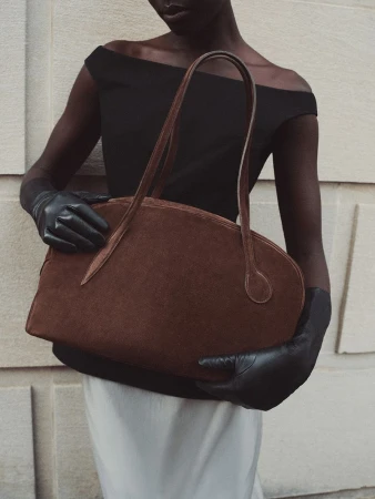 Model wearing a black off-the-shoulder top and black leather gloves holding a large brown suede handbag against a stone wall.