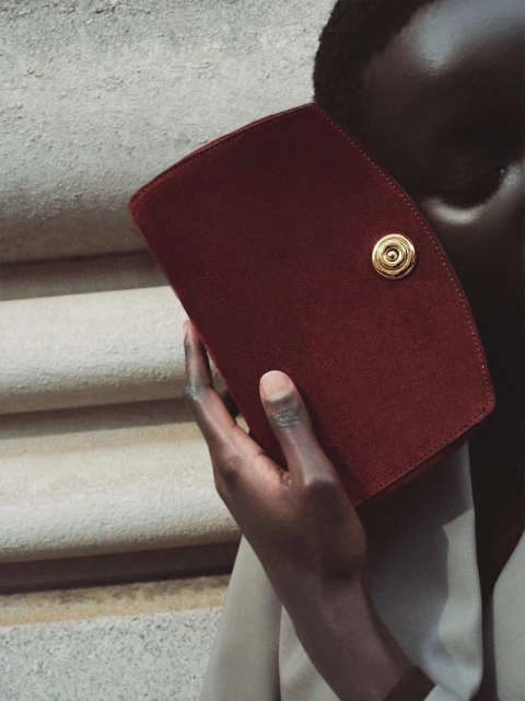 Dark-skinned adult holding a burgundy suede clutch with a gold clasp against a light beige background with a staircase and cloth.