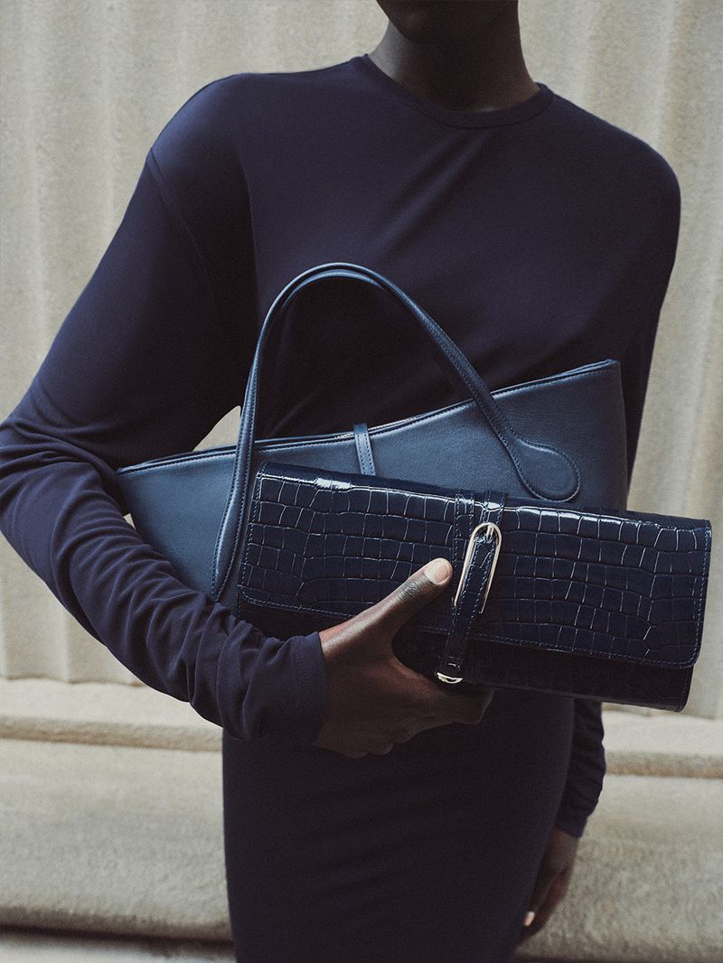 An African-American adult female carries two dark blue leather bags. The photo uses indoor lighting and a neutral wall background.