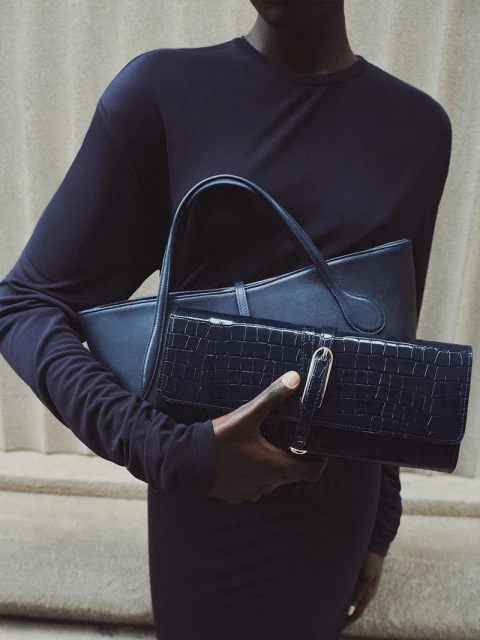 An African-American adult female carries two dark blue leather bags. The photo uses indoor lighting and a neutral wall background.