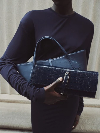 An African-American adult female carries two dark blue leather bags. The photo uses indoor lighting and a neutral wall background.