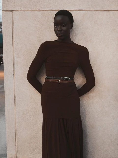 A slender African-American woman poses in a fitted brown dress and belt against a neutral wall in soft, natural light.