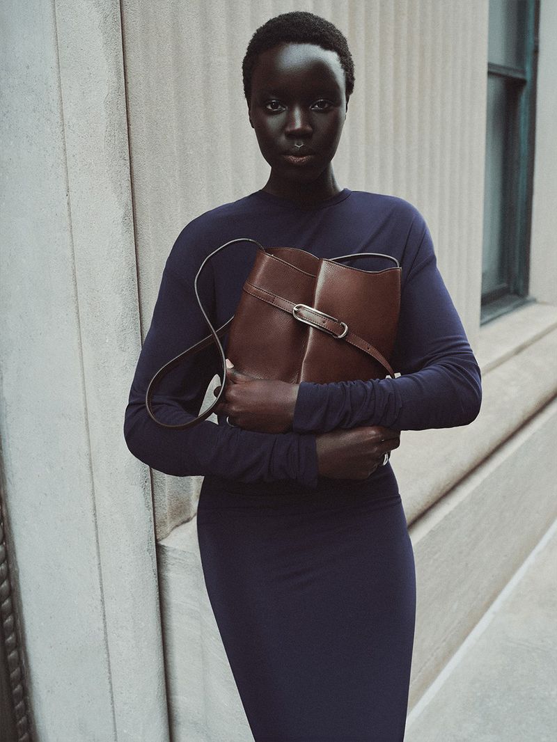 Black woman with short buzzcut hair wearing a navy blue long-sleeve dress and holding a brown leather bucket bag against a stone wall.