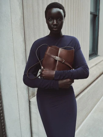 Black woman with short buzzcut hair wearing a navy blue long-sleeve dress and holding a brown leather bucket bag against a stone wall.