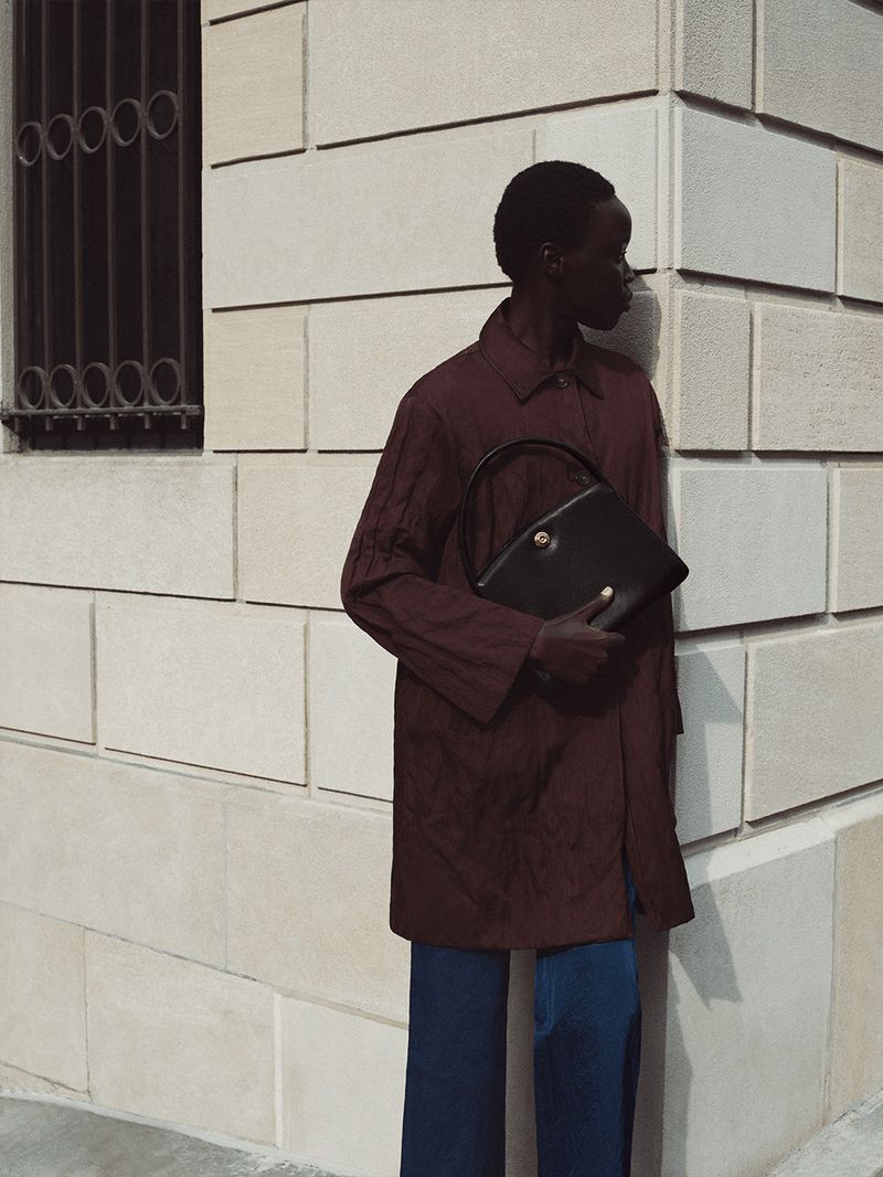 Black woman wearing a burgundy coat holding a black handbag against a stone architectural building wall