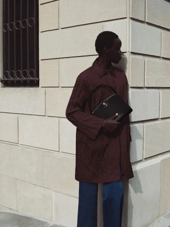 Black woman wearing a burgundy coat holding a black handbag against a stone architectural building wall