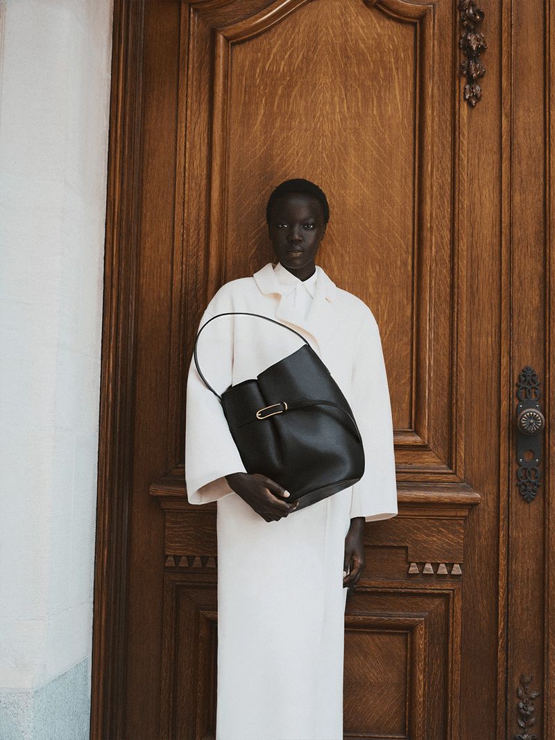 Fashion shot of a black leather hobo bag held by a woman in a white coat, standing in front of a paneled wood door. Minimalist aesthetic.