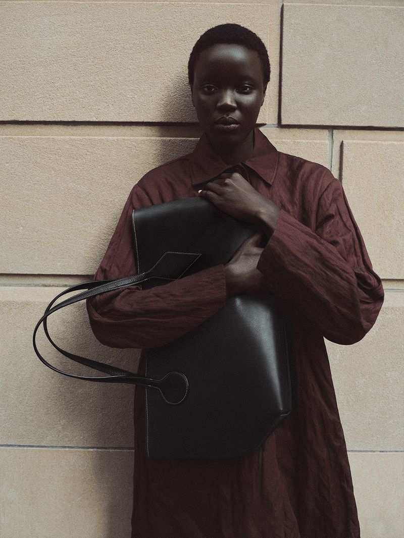 Adult female carrying a black leather tote bag in front of a tan-colored brick wall, wearing a textured long-sleeve dress.
