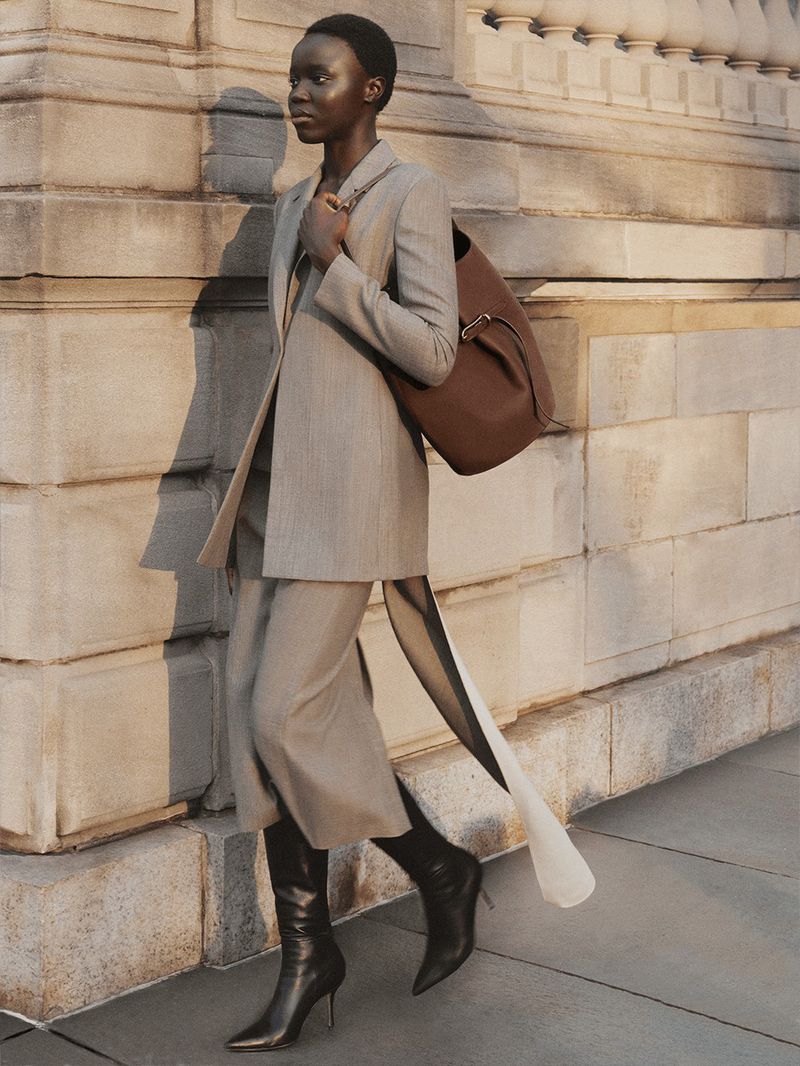 Black woman walking in a tailored grey suit and black boots carrying a large brown leather tote bag by a stone building facade.
