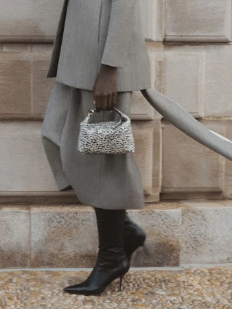 Cropped shot of a model in a grey tailored skirt suit and black stiletto boots holding a small animal print handbag.