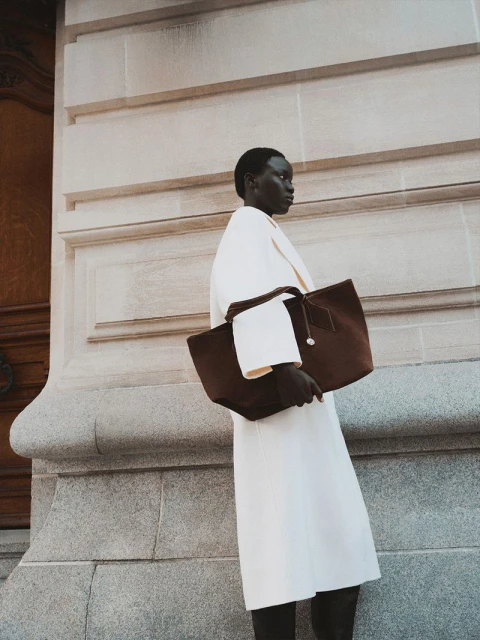 A dark-skinned woman wearing a light-colored coat holds a brown tote bag next to a large stone wall in an outdoor setting during the daytime.