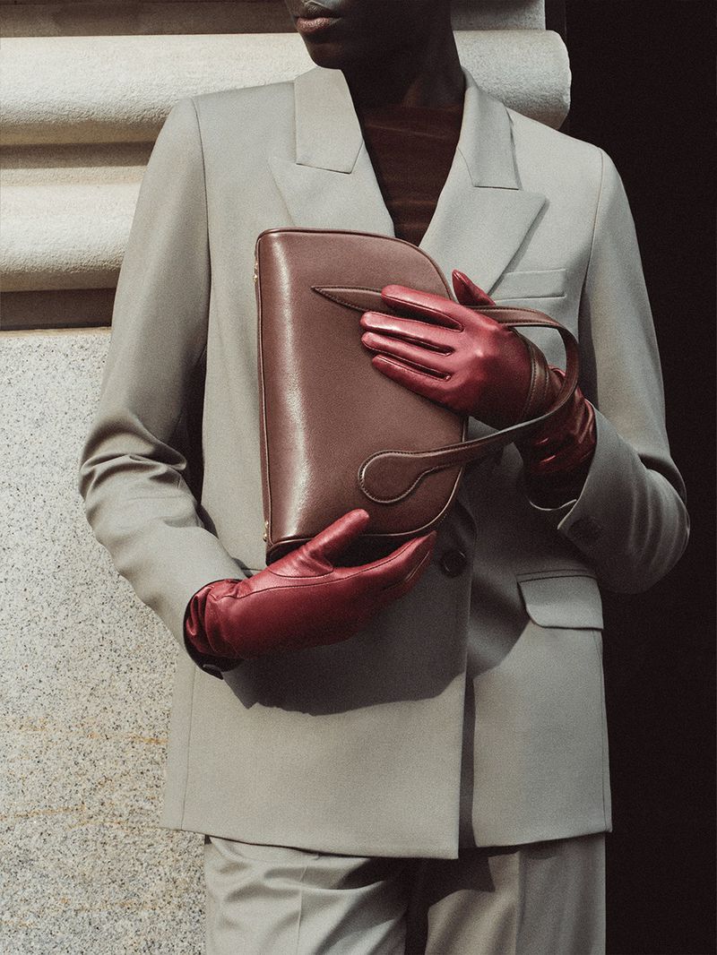 Close-up of a dark-skinned model holding a brown clutch, wearing matching gloves and a neutral suit against a textured wall.